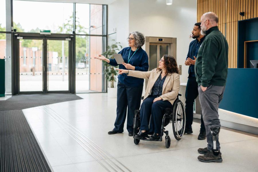 Occupational Therapy led accessibility consultant reviewing inclusive building access with a wheelchair user, mobility impaired person and facilities managers in a modern public sector reception area.